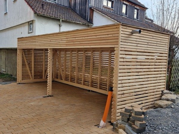 Doppelcarport aus hellem Holz mit Rhombusfassade an Seiten und Rückwand, davor eine gepflasterte Einfahrt.