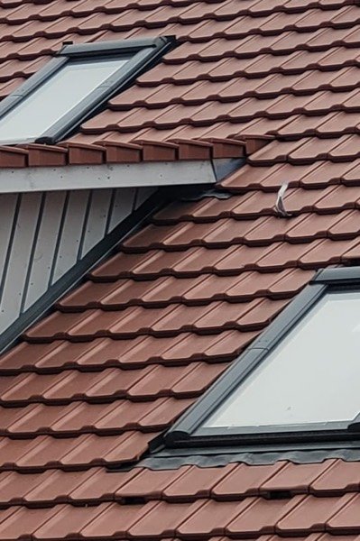 Ziegeldach mit zwei neu eingebauten Dachfenstern in dunklen Eindeckrahmen, daneben die grau verkleidete Seitenwand einer Gaube.