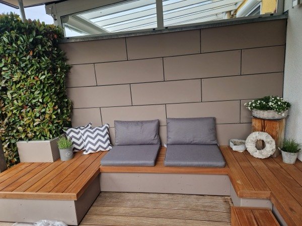 L-förmige Sitzlounge mit Sitzflächen aus rötlichem Holz und grauen Polstern vor einer Wand aus großformatigen Platten, links eine Hecke.